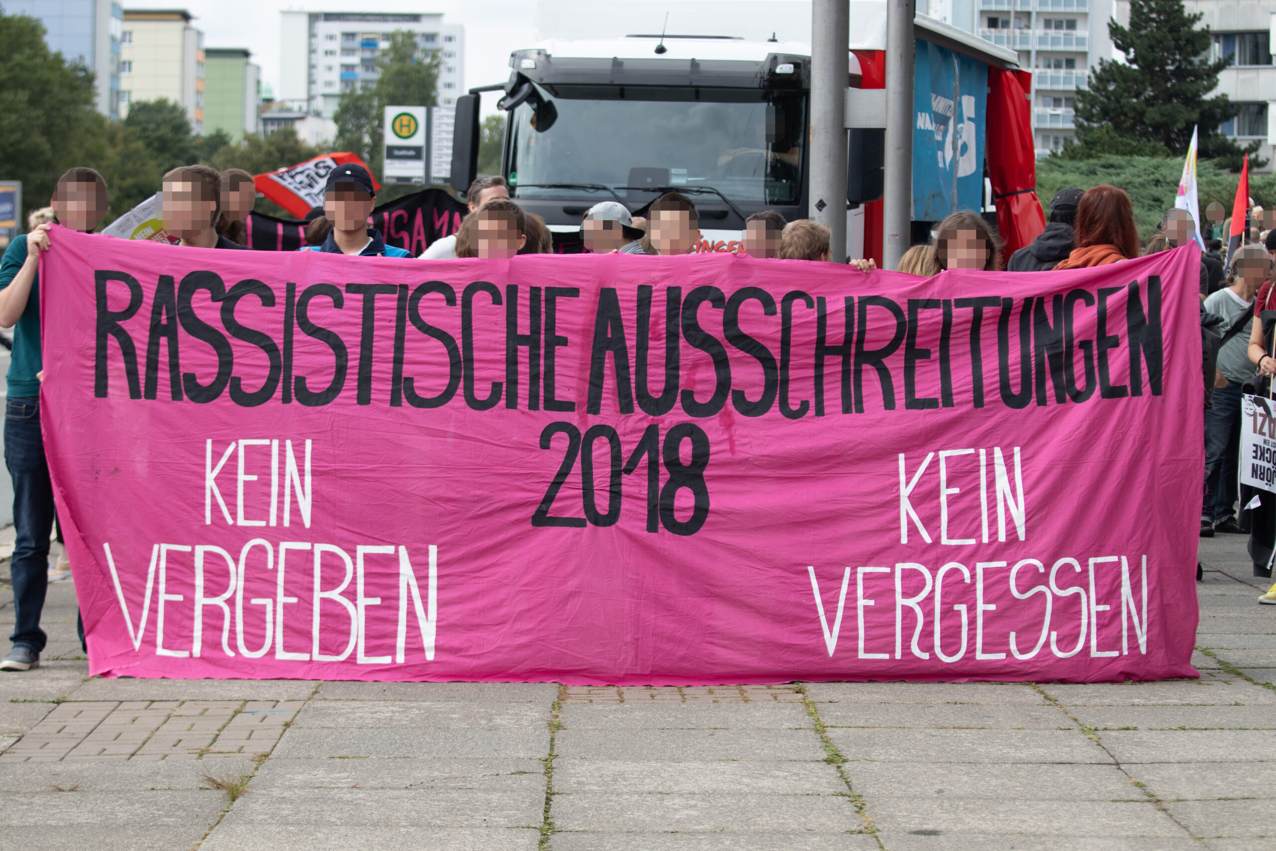 Demonstrationstransparent aus pinkem Stoff, dahinter Menschen und ein Lastwagen. Auf dem Transparent steht: 